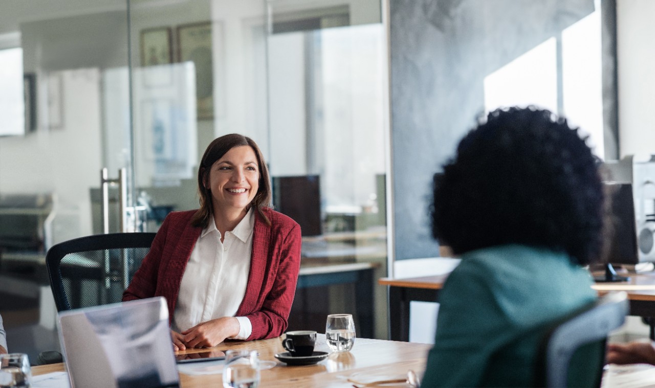 A woman in a red blazer is sitting at a conference table, smiling and looking at another woman.