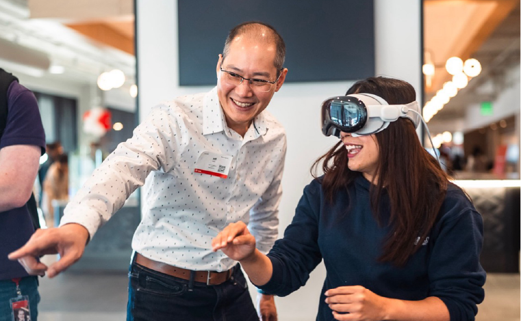 Image of two people, the one on the left is guiding a person who is wearing a virtual reality headset