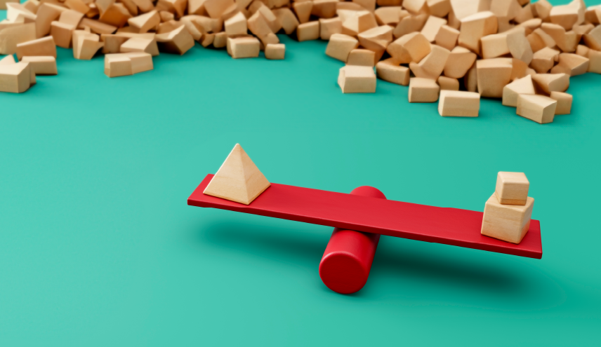 A balanced seesaw with a wooden pyramid on one side and two stacked wooden cubes on the other, illustrating a comparison of value. A large pile of additional wooden blocks is in the background.